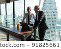 Picture of two business men negotiating over a deal. One man is in his 70th he is wearing black suit. The other man is in his 40th he is wearing a white shirt and black tie. They sat in lounge. 91825661