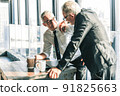 Picture of two business men negotiating over a deal. One man is in his 70th he is wearing black suit. The other man is in his 40th he is wearing a white shirt and black tie. They sat in lounge. 91825663