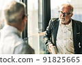 Picture of two business men negotiating over a deal. One man is in his 70th he is wearing black suit. The other man is in his 40th he is wearing a white shirt and black tie. They sat in lounge. 91825665