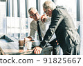 Picture of two business men negotiating over a deal. One man is in his 70th he is wearing black suit. The other man is in his 40th he is wearing a white shirt and black tie. They sat in lounge. 91825667