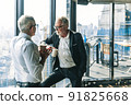 Picture of two business men negotiating over a deal. One man is in his 70th he is wearing black suit. The other man is in his 40th he is wearing a white shirt and black tie. They sat in lounge. 91825668