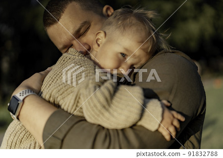 young dad with eyes closed hugging baby son putting head to his shoulder and sucking a pacifier. 91832788