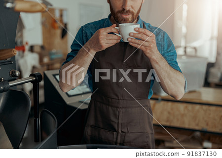 Close up of male barista hands holding cup of coffee on background of roasting coffee machine Close up of male barista hands holding cup of coffee on background of roasting coffee machine 91837130