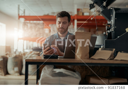 Smiling business owner of small coffee roasting factory working on workplace with laptop and phone Smiling business owner of small coffee roasting factory working on workplace with laptop and phone 91837168