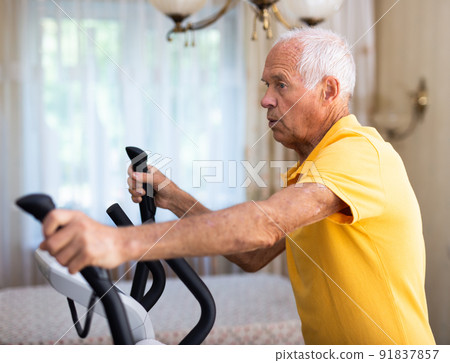 Elderly man exercising on elliptic trainer in living room 91837857