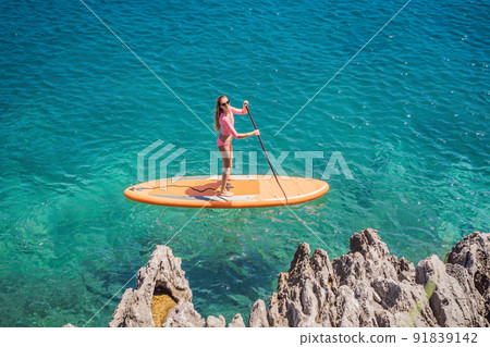 Young women Having Fun Stand Up Paddling in blue water sea in Montenegro. SUP. girl Training on Paddle Board near the rocks 91839142