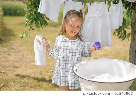 Girl holds bottle of liquid laundry detergent in hands, standing at basin for hand washing clothes. Snow-white baby underwear on clothesline. Advertising of eco friendly hypoallergenic detergents 91843908