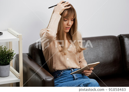 Upset confused unhappy blonde student lady with copybook touch head sitting in sofa couch at modern living room interior. Female client have shrink appointment in psychological help office. Copy space 91858483