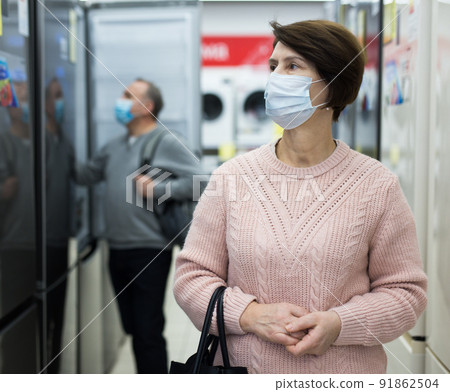Middle aged woman in mask choosing refrigerator in appliance store Middle aged woman in mask choosing refrigerator in appliance store 91862504