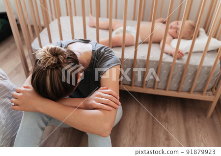 Crying mother sitting next to baby crib with head on her knees suffering postnatal depression Crying mother sitting next to baby crib with head on her knees suffering postnatal depression 91879923