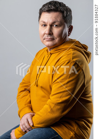 Portrait of confident handsome Caucasian man looking at camera, sitting on chair, looks calm, wearing urban style hoodie. Indoor studio shot isolated on gray background. 91884327