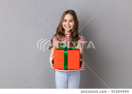 Portrait of smiling little girl wearing striped T-shirt giving present and congratulating on birthday, mother's day, offering gift box for charity. Indoor studio shot isolated on gray background. Portrait of smiling little girl wearing striped T-shirt giving present and congratulating on birthday, mother's day, offering gift box for charity. Indoor studio shot isolated on gray background. 91884404