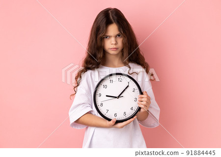 Portrait of little girl wearing white T-shirt holding wall clock, being unhappy, deadline, being sad, not finished her task in time. Indoor studio shot isolated on pink background. 91884445