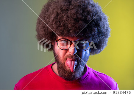 Portrait of hipster man with Afro hairstyle showing out tongue with naughty disobedient grimace, making face, wearing red sweatshirt. Indoor studio shot isolated on colorful neon light background. 91884476