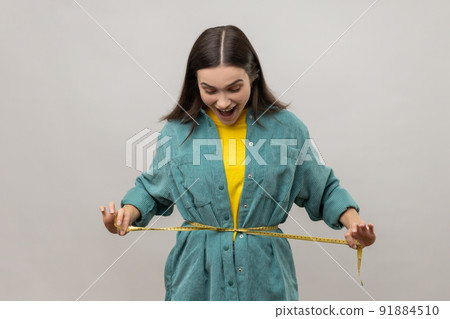 Smiling positive woman holding tape line on her waist, being amazed with her body parameters after slimming, wearing casual style jacket. Indoor studio shot isolated on gray background. 91884510
