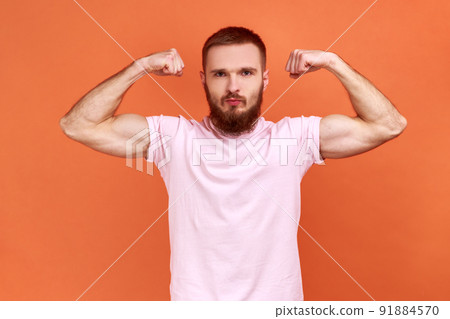Portrait of strong bearded man demonstrating power in his hands, showing biceps, feeling energy to win success, wearing pink T-shirt. Indoor studio shot isolated on orange background. 91884570