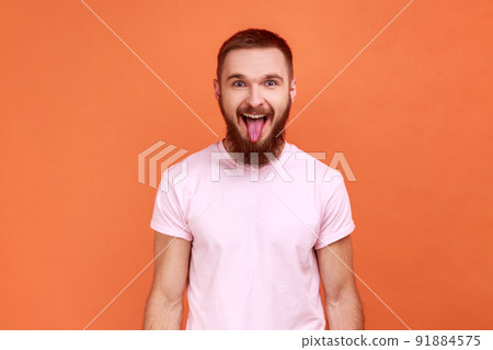 Portrait of bearded man showing out tongue and looking at camera with naughty disobedient grimace, making face, wearing pink T-shirt. Indoor studio shot isolated on orange background. 91884575