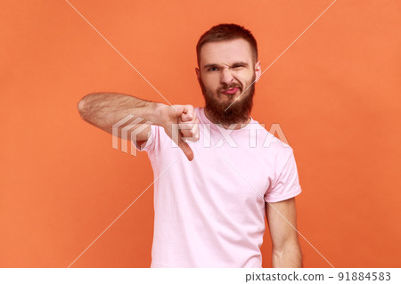 Portrait of man criticizing bad quality with thumbs down displeased grimace, showing dislike gesture, expressing disapproval, wearing pink T-shirt. Indoor studio shot isolated on orange background. Portrait of man criticizing bad quality with thumbs down displeased grimace, showing dislike gesture, expressing disapproval, wearing pink T-shirt. Indoor studio shot isolated on orange background. 91884583