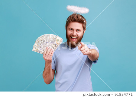 Portrait of satisfied bearded angelic man with holy nimbus over head showing fan of money, holding dollar bills and pointing to camera with index finger. Indoor studio shot isolated on blue background 91884771
