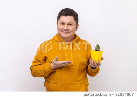 Portrait of satisfied positive man holding presenting yellow flower pot and cactus, looking at camera with toothy smile, wearing urban style hoodie. Indoor studio shot isolated on white background. Portrait of satisfied positive man holding presenting yellow flower pot and cactus, looking at camera with toothy smile, wearing urban style hoodie. Indoor studio shot isolated on white background. 91884807