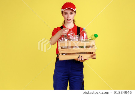Portrait of serious worker woman standing with box of empty plastic bottles and pointing at camera, choosing you, wearing overalls and red cap. Indoor studio shot isolated on yellow background. 91884946