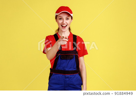 Portrait of excited happy worker woman standing and pointing at camera, laughing loud out, hearing funny joke, wearing overalls and red cap. Indoor studio shot isolated on yellow background. Portrait of excited happy worker woman standing and pointing at camera, laughing loud out, hearing funny joke, wearing overalls and red cap. Indoor studio shot isolated on yellow background. 91884949