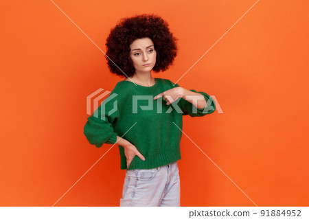 Portrait of selfish woman with Afro hairstyle wearing green casual style sweater pointing at herself with finger, having proud expression. Indoor studio shot isolated on orange background. 91884952