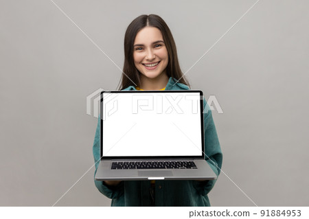 Satisfied delightful woman standing showing laptop with white empty screen for promotion, looking at camera with smile, wearing casual style jacket. Indoor studio shot isolated on gray background. Satisfied delightful woman standing showing laptop with white empty screen for promotion, looking at camera with smile, wearing casual style jacket. Indoor studio shot isolated on gray background. 91884953