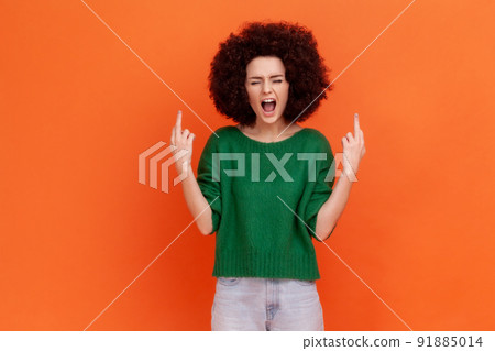 Angry aggressive woman with Afro hairstyle wearing green casual style sweater screaming angrily, showing middle finger with both hands. Indoor studio shot isolated on orange background. 91885014