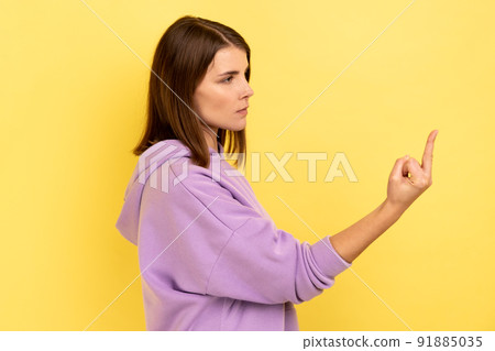 Side view portrait of rude vulgar woman showing middle fingers, impolite gesture of disrespect, looking with aggression hate, wearing purple hoodie. Indoor studio shot isolated on yellow background. 91885035