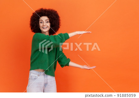 Positive woman with Afro hairstyle wearing green casual style sweater showing big size with both hands, looking at camera with happy expression. Indoor studio shot isolated on orange background. Positive woman with Afro hairstyle wearing green casual style sweater showing big size with both hands, looking at camera with happy expression. Indoor studio shot isolated on orange background. 91885059