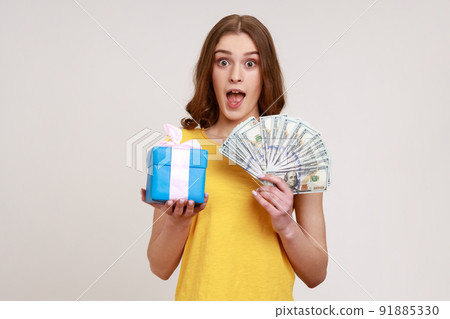 Wow, holiday present shopping. Portrait of amazed teen girl in yellow T-shirt showing dollar banknotes, cashback money and gift box, looking shocked. Indoor studio shot isolated on gray background. Wow, holiday present shopping. Portrait of amazed teen girl in yellow T-shirt showing dollar banknotes, cashback money and gift box, looking shocked. Indoor studio shot isolated on gray background. 91885330
