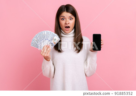 Shocked female holds big sum of dollars and showing smart phone with blank screen for advertisement, wearing white casual style sweater. Indoor studio shot isolated on pink background. Shocked female holds big sum of dollars and showing smart phone with blank screen for advertisement, wearing white casual style sweater. Indoor studio shot isolated on pink background. 91885352