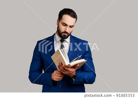 Concentrated bearded man reading book, enjoying reading, standing absorbed with exciting plot, checking schedule in notepad, wearing official style suit. Indoor studio shot isolated on gray background Concentrated bearded man reading book, enjoying reading, standing absorbed with exciting plot, checking schedule in notepad, wearing official style suit. Indoor studio shot isolated on gray background 91885353
