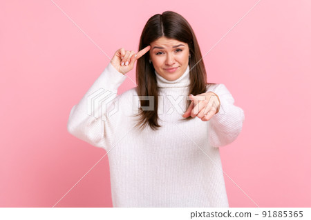 Portrait of beautiful woman holding finger near head and pointing finger at camera, stupid gesture, wearing white casual style sweater. Indoor studio shot isolated on pink background. Portrait of beautiful woman holding finger near head and pointing finger at camera, stupid gesture, wearing white casual style sweater. Indoor studio shot isolated on pink background. 91885365