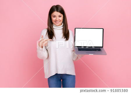 Good looking female with dark hair holding laptop in hands, pointing finger at empty display for ad, wearing white casual style sweater. Indoor studio shot isolated on pink background. 91885380