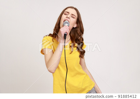 Portrait of excited positive teenager girl with brown hair in yellow casual t-shirt singing songs at karaoke holding microphone, entertainment. Indoor studio shot isolated on gray background. 91885412