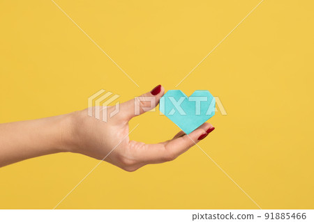 Profile side view closeup of woman hand holding tiny turquoise heart, symbol of love, showing romantic feelings. Indoor studio shot isolated on yellow background. 91885466