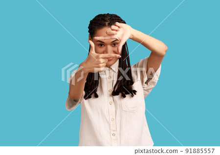 Happy ambitious woman with black dreadlocks makes hand frames, searches perfect angle, smiling, gazes at camera through hands, wearing white shirt. Indoor studio shot isolated on blue background. Happy ambitious woman with black dreadlocks makes hand frames, searches perfect angle, smiling, gazes at camera through hands, wearing white shirt. Indoor studio shot isolated on blue background. 91885557