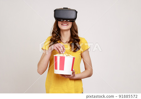 Young smiling female in VR headset watching movie with popcorn, smiling happily, expressing positive emotions, wearing yellow T-shirt. Indoor studio shot isolated on gray background. 91885572