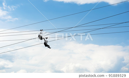 Zipline riders, silhouettes of two people on blue sky background Zipline riders, silhouettes of two people on blue sky background 91889329