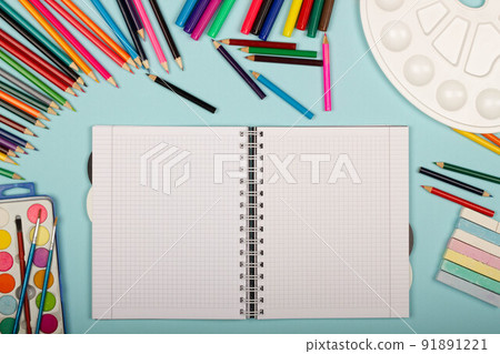 Blank sheet of paper of an open math notebook, art school supplies for painting, on a blue background with copy space for text. Colorful pencils, markers, paints, crayons. Top view. Back to school 91891221