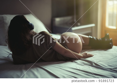 Beautiful young naked woman in headphones listening music lying on a bed in the bedroom near window at sunset. 91891723