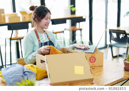 Portrait of a small startup business, SME owner, Asian female entrepreneur, talks to via video conferance, lets customers see products before pricing and packing them into boxes. Online Business. 91892853
