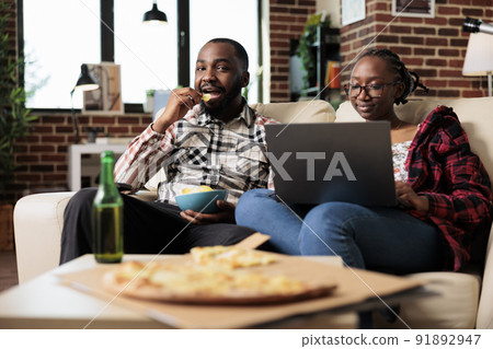 African american couple eating chips and using laptop in front of television, watching movie on channel program. Enjoying leisure activity with computer and takeaway fast food delivery. 91892947