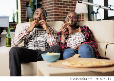 Young man and woman eating slice of pizza and drinking bottles of beer while they watch movie on tv together. Enjoying fast food takeaway delivery and beverage, watching television. Young man and woman eating slice of pizza and drinking bottles of beer while they watch movie on tv together. Enjoying fast food takeaway delivery and beverage, watching television. 91893215
