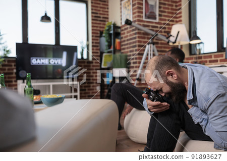 Caucasian man losing at shooting video games strategy, feeling frustrated and angry about lost competition game. Sad person being irritated after shooter gaming lose at gathering with friends. 91893627