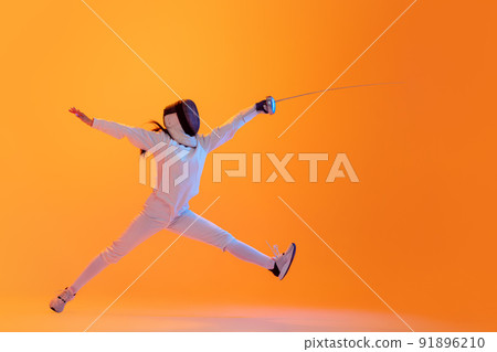Studio shot of professional fencer in white fencing costume and mask in action, motion isolated on orange color background. Sport, youth, activity, skills, 91896210