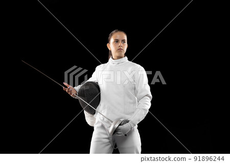 Young serious girl, beginner fencer in white fencing costume standing with rapier ans mask isolated on dark background. Sport, youth, achievements, challenges. 91896244