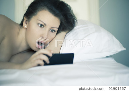 A girl lies in bed under the sheet can't fall asleep looking on mobile phone. Insomnia. Psychology. A girl lies in bed under the sheet can't fall asleep looking on mobile phone. Insomnia. Psychology. 91901367